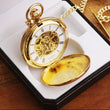 Load image into Gallery viewer, A gold-plated, skeleton double hunter pocket watch with a white Roman dial, displayed inside a luxury presentation box next to a matching Albert chain and a small piece of yellow jewelry, all set on a wooden surface.